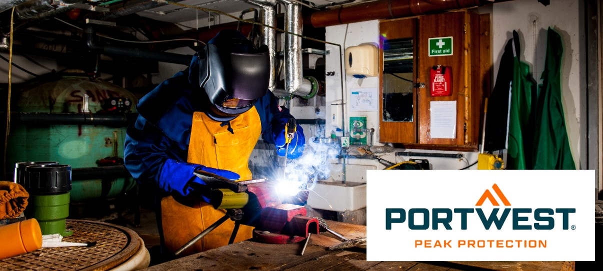 A worker is wearing a protective mask and flame-retardant protective equipment in an industrial environment. He is carrying out welding work and is holding a welding torch. In the background, pipes, tools and a first aid kit can be seen. In the bottom right of the image is the logo of "Portwest Peak Protection".