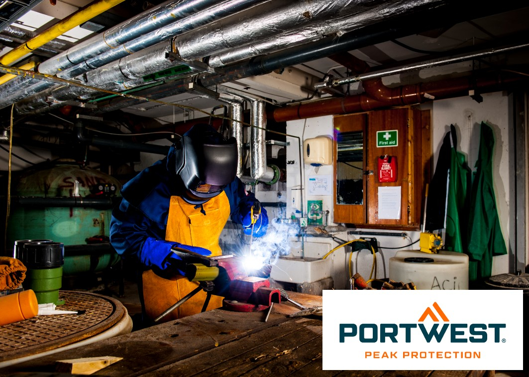 A worker is wearing a protective mask and flame-retardant protective equipment in an industrial environment. He is carrying out welding work and is holding a welding torch. In the background, pipes, tools and a first aid kit can be seen. In the bottom right of the image is the logo of "Portwest Peak Protection". There is a link that leads to our collection of flame-retardant clothing.