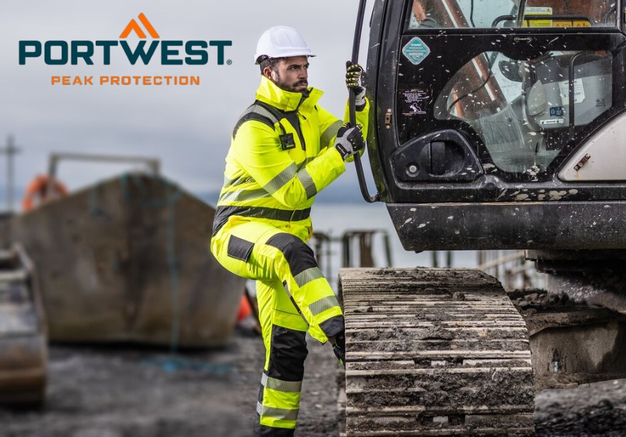 A worker in a bright yellow high-visibility suit with reflective stripes, a white safety helmet and gloves climbs into a construction machine. In the background you can see a construction site environment with construction equipment and a gray sky. At the top left of the picture is the "Portwest" logo with the slogan "Peak Protection". A link to our selection of different work overalls is provided.