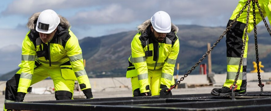 Three workers in yellow winter clothing and white helmets in front of a mountain backdrop with a blue, slightly cloudy sky.