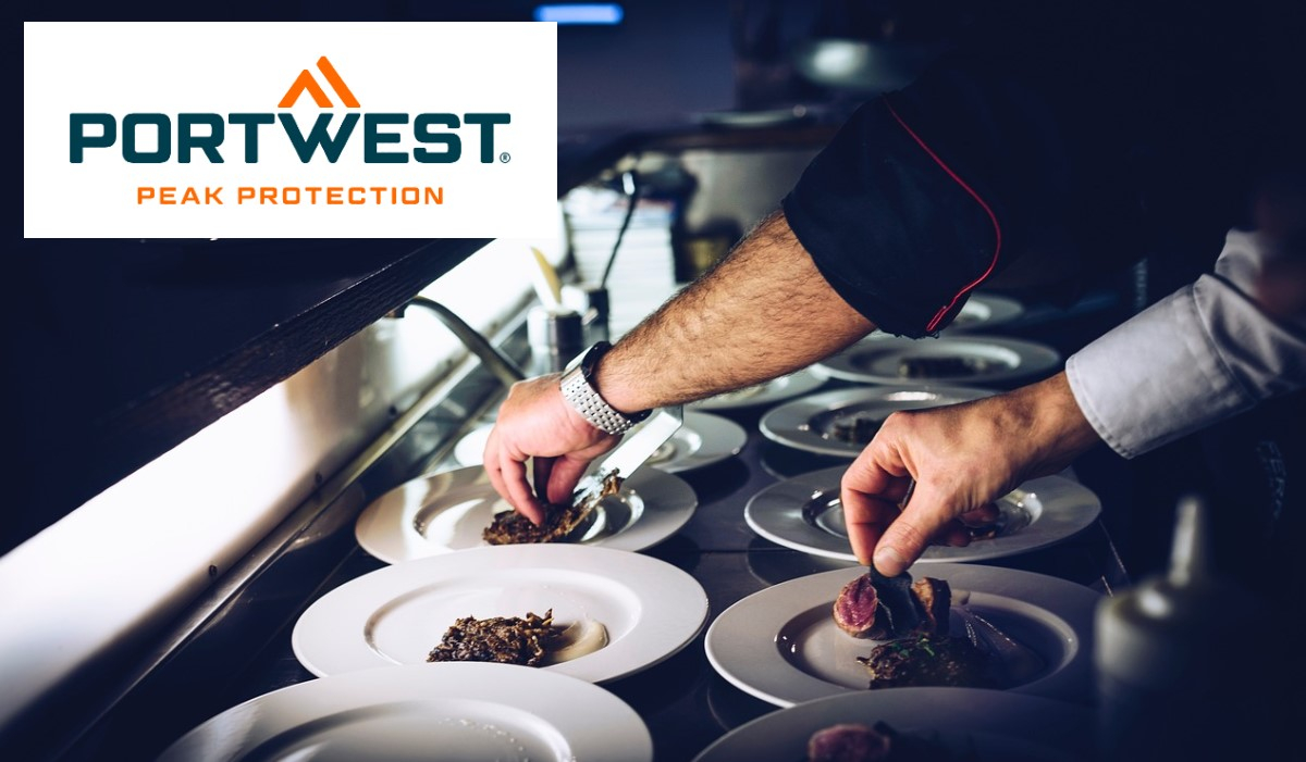 Two chefs, of whom only one arm can be seen in a black chef's jacket and one in a white chef's jacket, carefully arrange food on white plates in a professional commercial kitchen. The Portwest "Peak Protection" logo is displayed at the top left. There is a link to our chef's clothing.