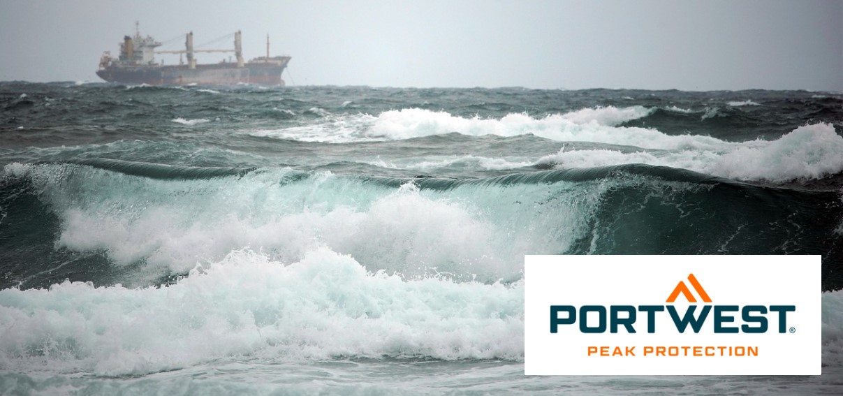 A cargo ship in the distance navigates through a stormy sea with high, foam-capped waves. In the foreground you can see strong, turbulent waves. The Portwest logo is in the bottom right of the picture.