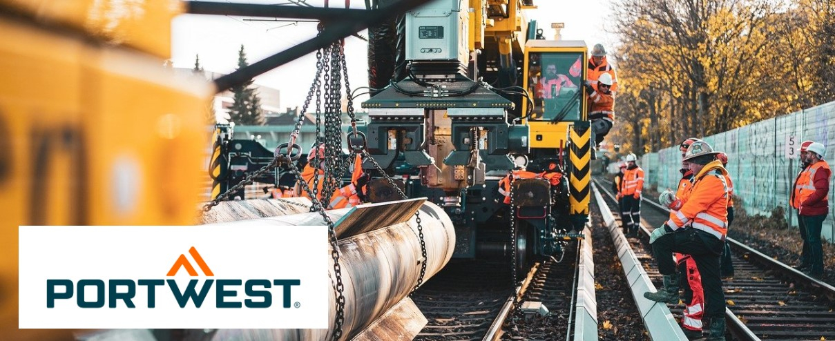 Several workers in orange high visibility clothing and safety helmets are carrying out construction work on a rail system. In the background you can see heavy equipment such as a crane and rail vehicle. Portwest logo visible in the corner.