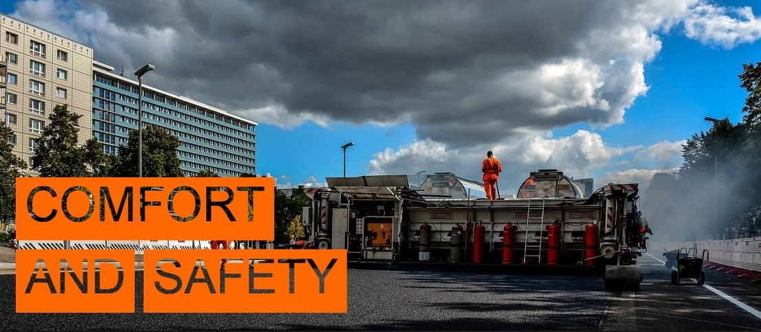 Scene of a road construction site. On the left of the picture you can see high-rise buildings and some trees, the right part of the picture is covered by a water fountain. In the bottom left of the picture the words "Comfort and Safety" are written in cut-out letters with an orange background.