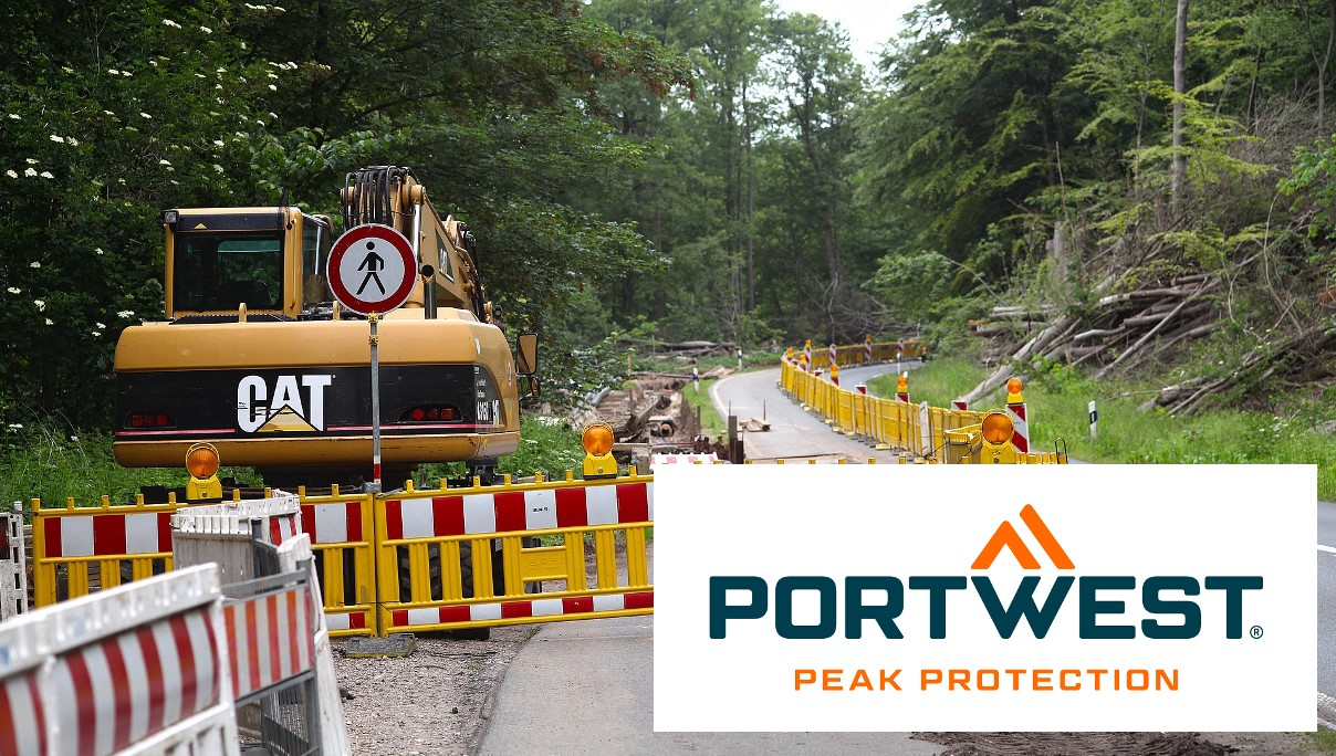 Construction site on a road in the forest. A yellow barrier marks the construction site. In the bottom right of the picture you can see the Portwest logo in blue and orange on a white background. There is a link that leads to our road construction protective clothing department.