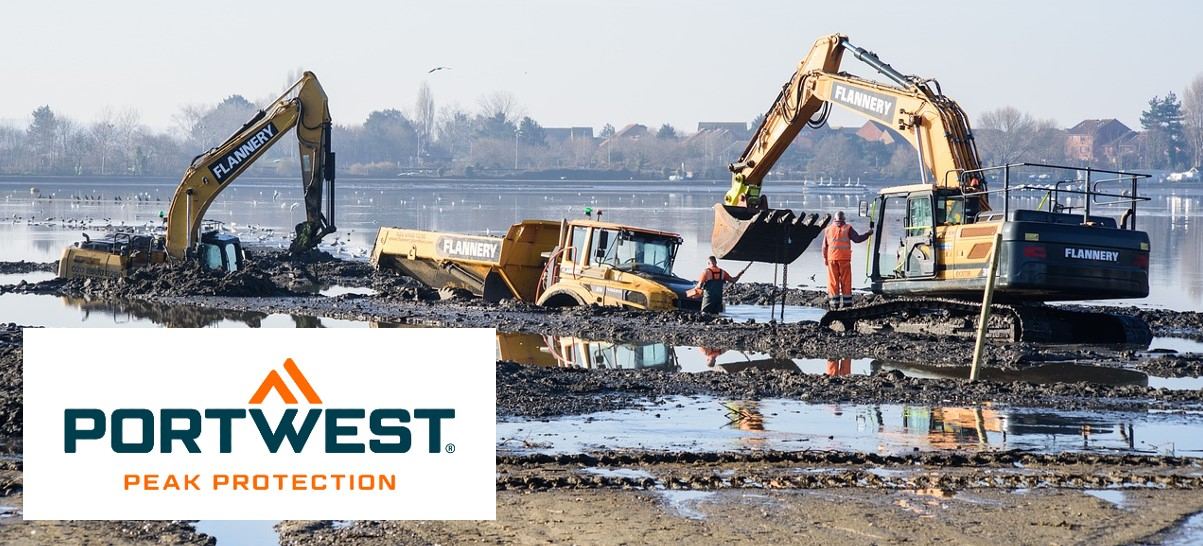 Construction site scene with excavators and large machines. There are puddles and mud on the ground, the sky is blue and you can see some houses and trees on the horizon.