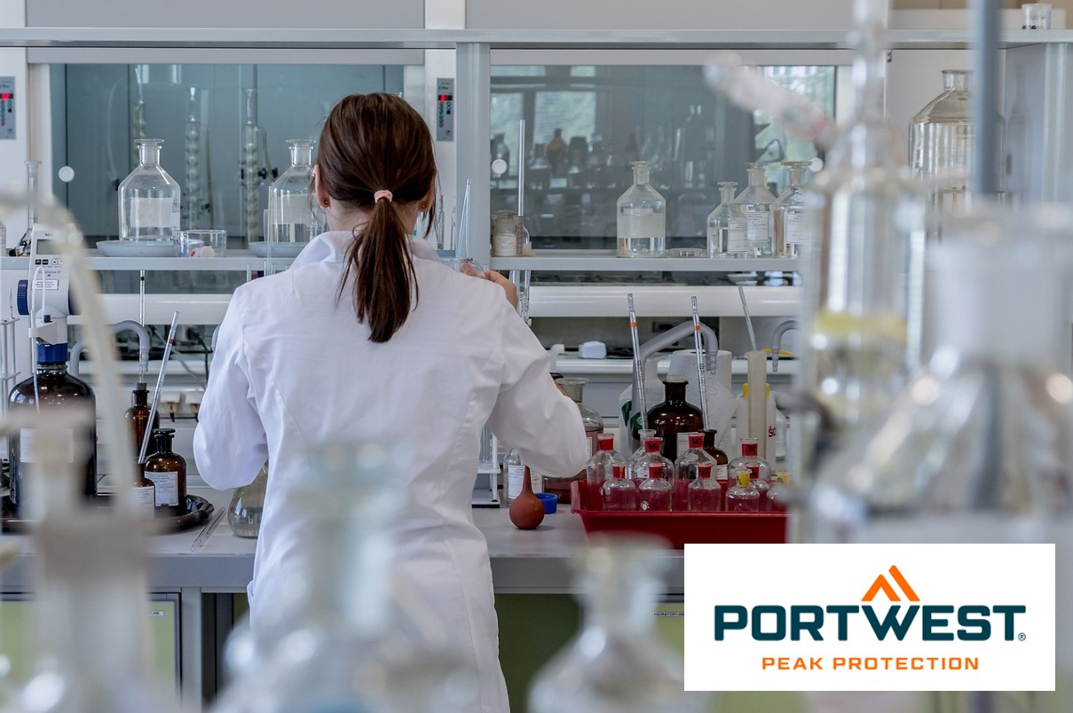 A woman with dark brown hair tied back wears a white lab coat and works in a modern laboratory with various laboratory equipment and chemical bottles. The logo of "Portwest Peak Protection" is at the bottom right of the picture. There is a link that leads to all our lab coats.