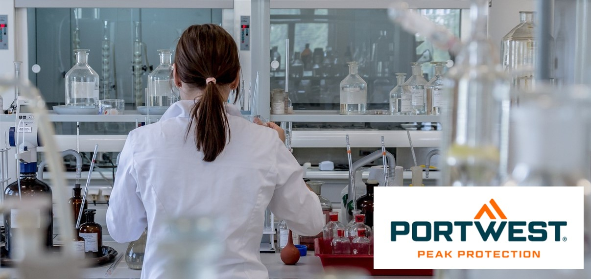A woman with dark brown hair tied back wears a white lab coat and works in a modern laboratory with various laboratory equipment and chemical bottles. The logo of "Portwest Peak Protection" is at the bottom right of the picture.