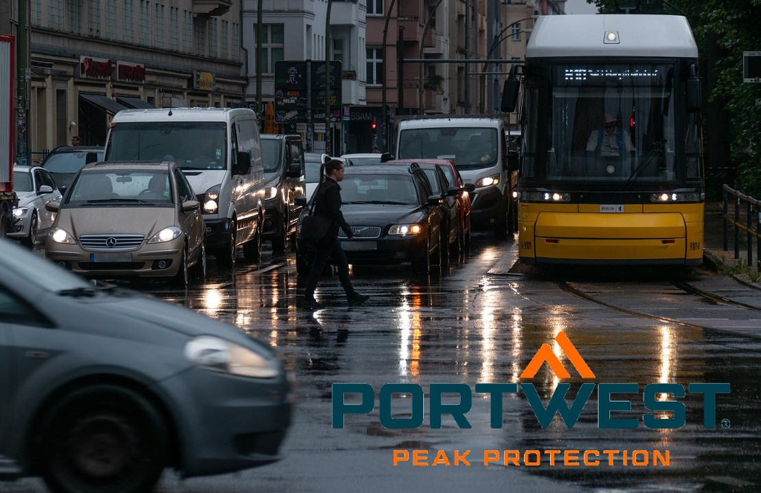 Street scene in the rain. Numerous cars and buses are parked at a crossroads with their headlights on. The Portwest logo in orange and blue is in the bottom right edge of the picture. There is a link that leads to our rain jackets.