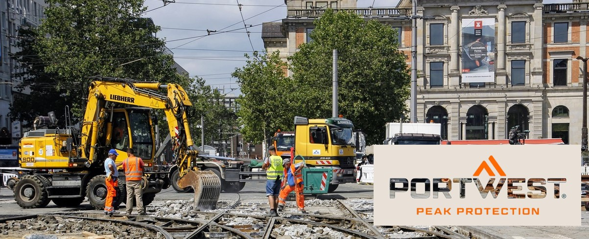 Construction workers in orange safety clothing work on a construction site in the city. In the background you can see excavators, trucks and a historic building. The Portwest logo and the slogan "Peak Protection" are shown at the bottom of the image.