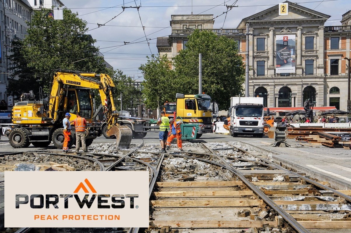 Construction workers in orange safety clothing work on a construction site in the city. In the background you can see excavators, trucks and a historic building. The Portwest logo and the slogan "Peak Protection" are shown at the bottom of the image. There is a link that leads to the article page for the cooling cap.